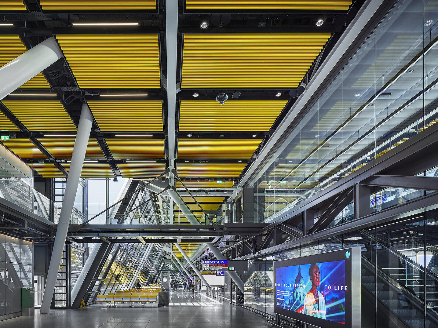Ein moderner, geräumiger Bahnhofsinnenraum mit gelben Lamellendecken, Glaswänden, Stahlträgern und Rolltreppen. Ein großer digitaler Bildschirm zeigt eine Werbung in der Nähe eines Glasgeländers und einer Treppe.