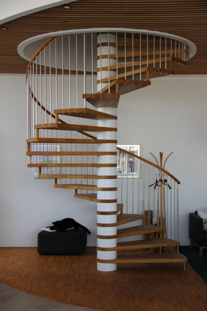 Eine moderne Wendeltreppe mit Holzstufen und Metallgeländer windet sich um eine weiße Mittelsäule. Eine schwarze Katze liegt auf einer dunklen Ottomane unter der Treppe, und in der Nähe steht ein hölzerner Kleiderständer.