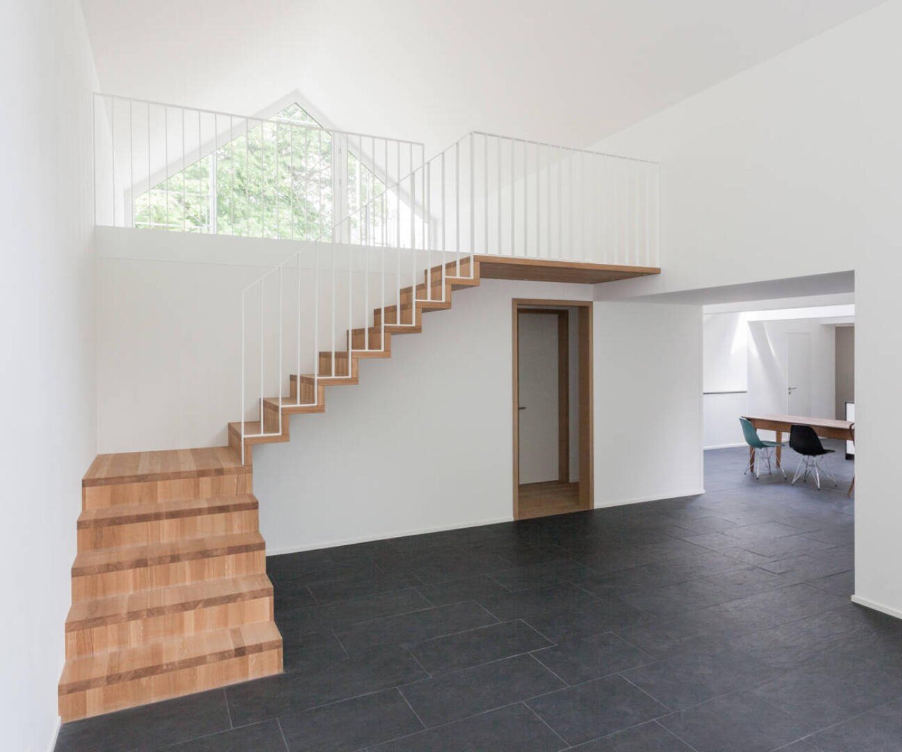 Intérieur minimaliste avec escalier en bois et rampe en métal blanc, carrelage noir, murs blancs et grandes fenêtres. Un petit espace de réunion avec chaises et table est visible en arrière-plan.