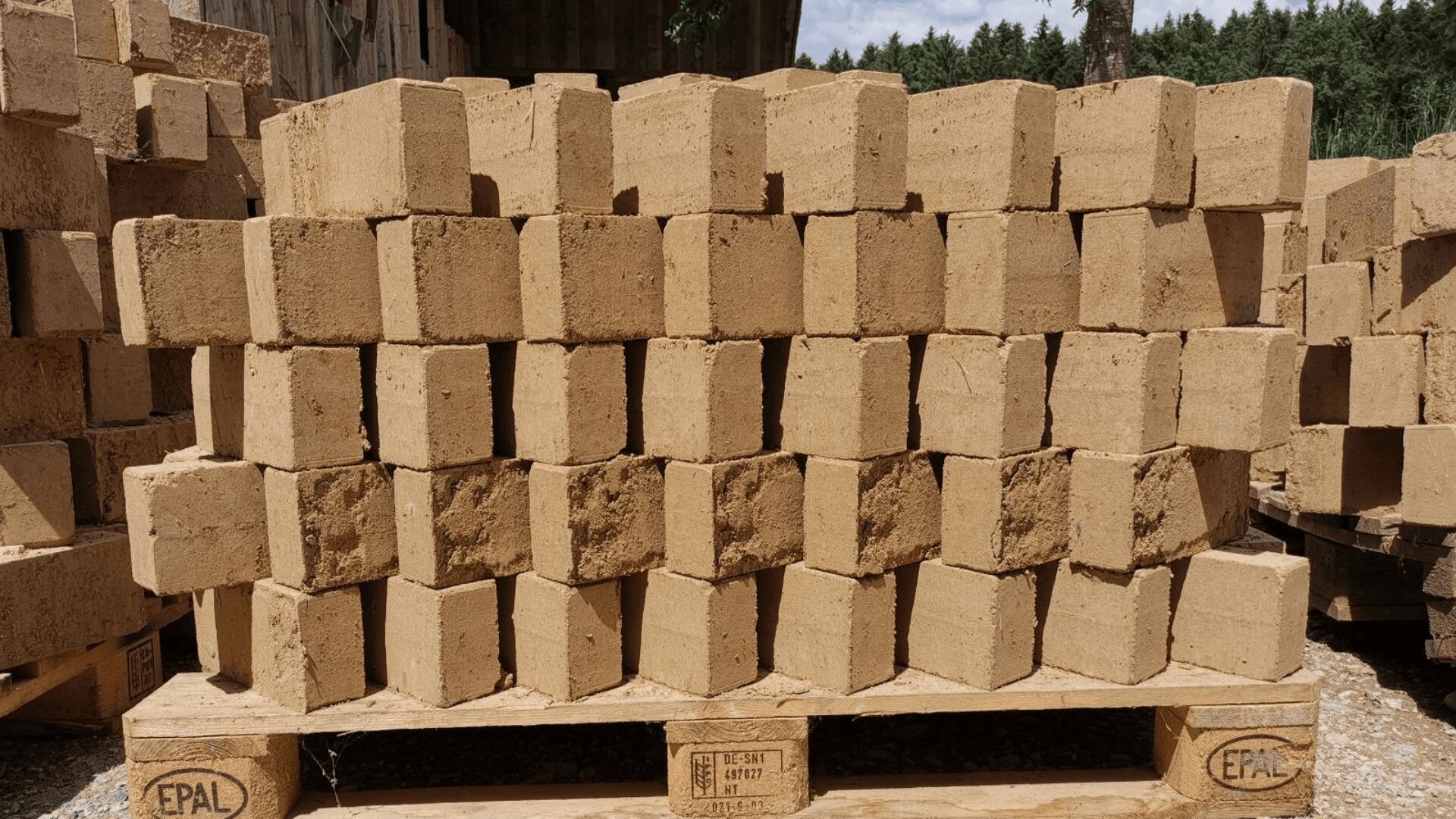 Ein Stapel rechteckiger Lehmziegel, die in Reihen auf einer Holzpalette im Freien angeordnet sind, wobei im Hintergrund weitere Ziegel und Bäume zu sehen sind.
