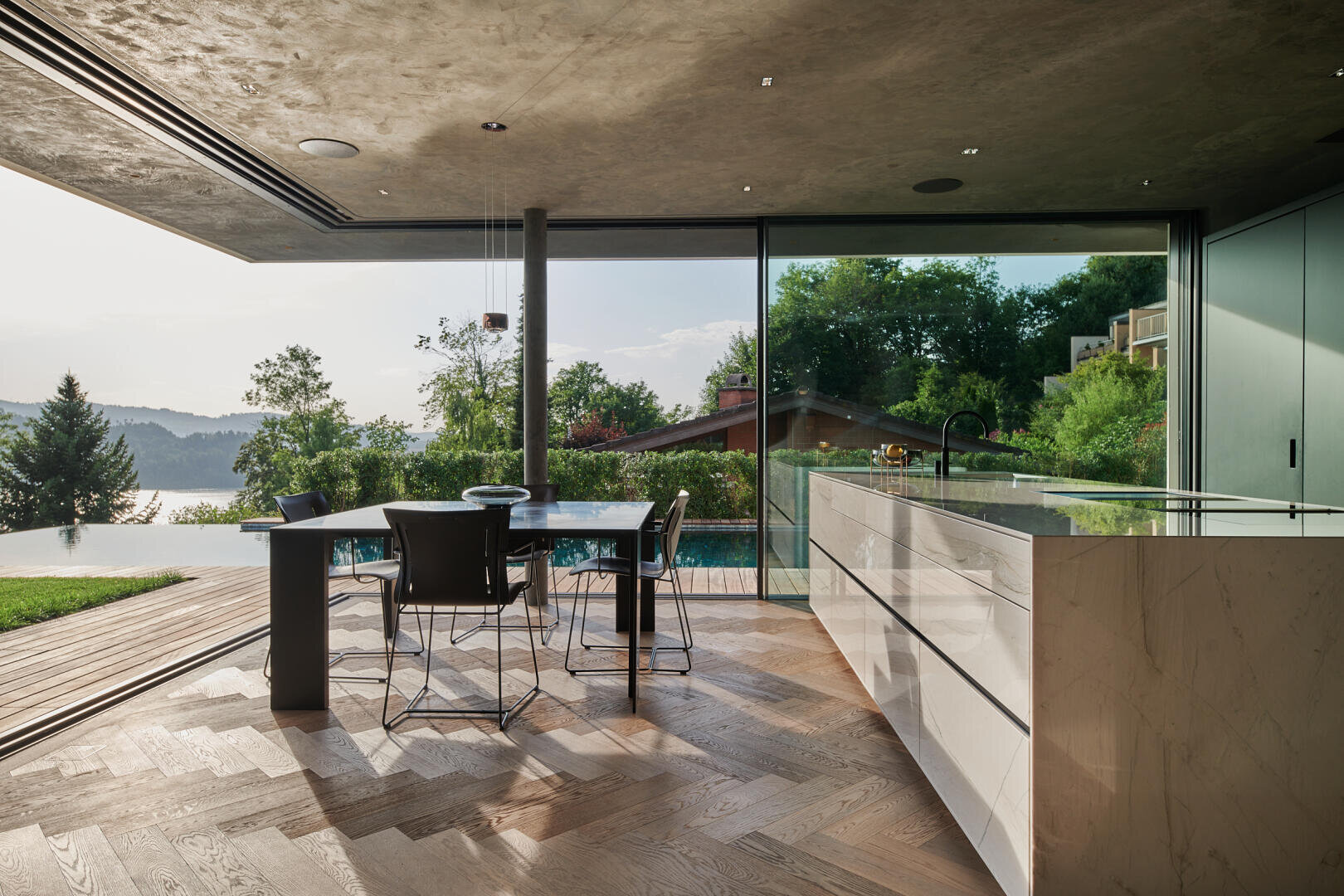 Moderne offene Küche und Essbereich mit Marmorarbeitsflächen und Holzböden, die sich zu einer Terrasse mit Pool, üppigem Grün und Blick auf einen See und Bäume im Hintergrund öffnen.