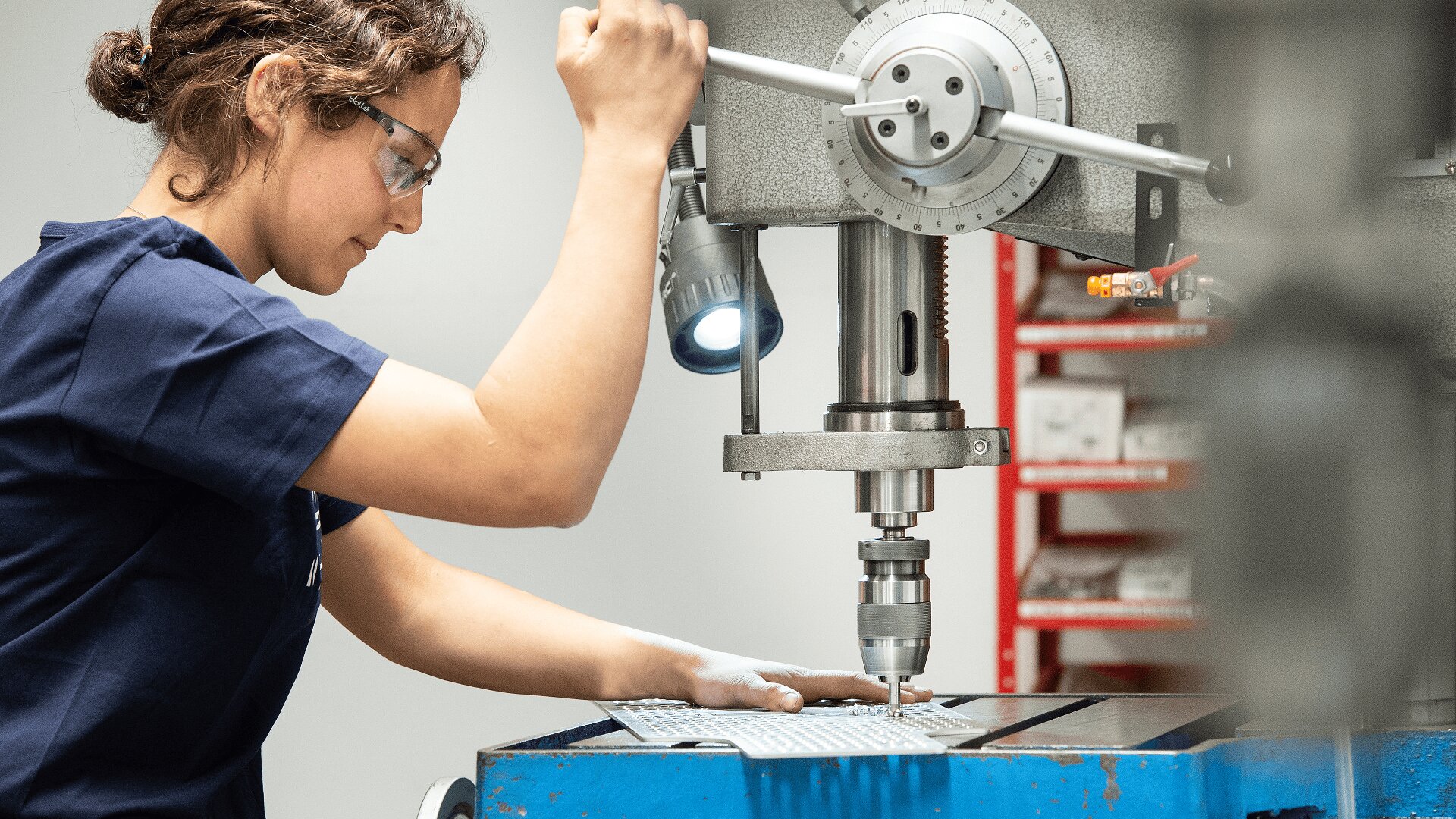 Eine Person, die eine Schutzbrille trägt, bedient eine große industrielle Bohrmaschine und führt ein Blech auf dem Arbeitstisch bei heller Beleuchtung in einer Werkstatt.