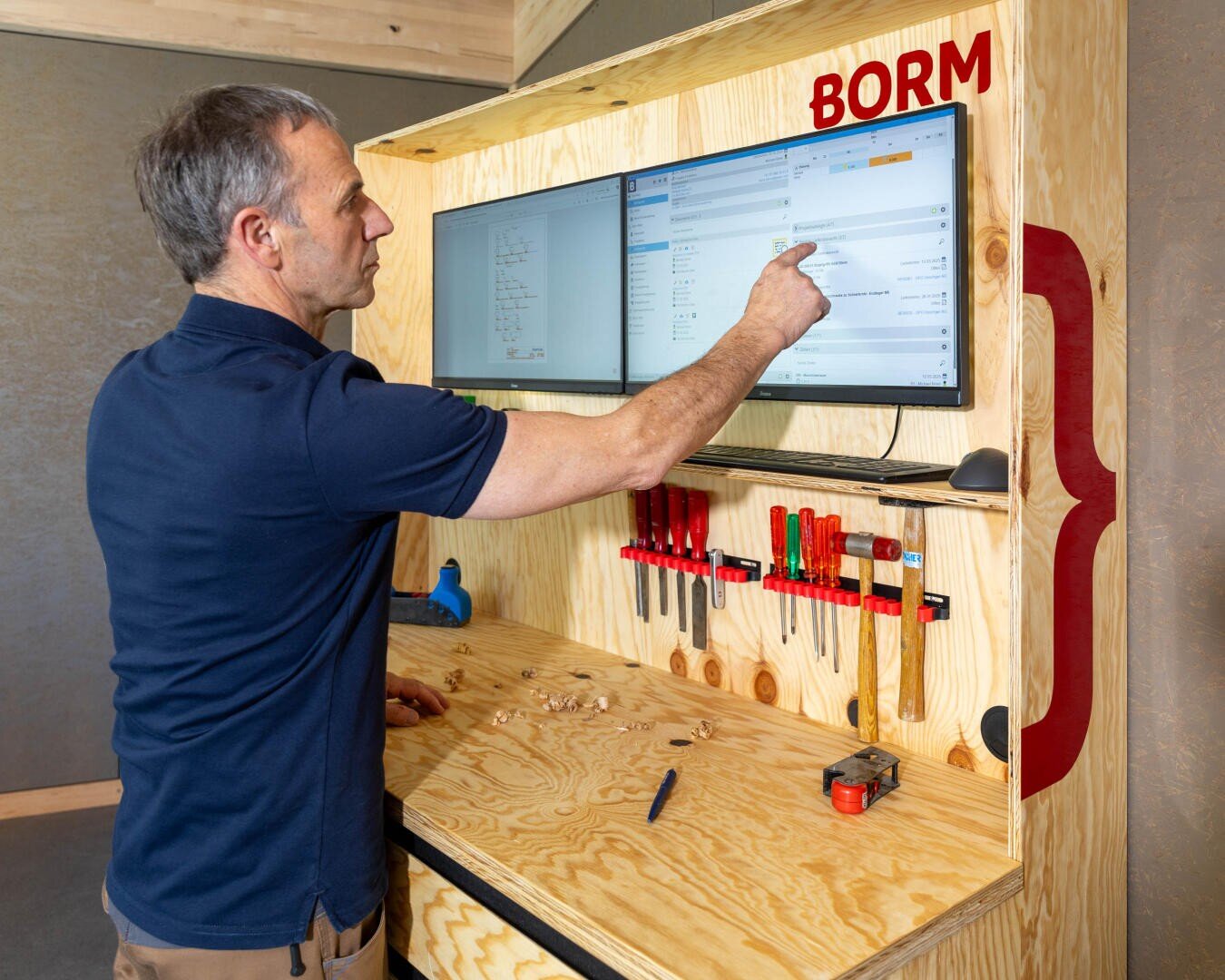 Un homme vêtu d'une chemise bleu marine utilise un moniteur à écran tactile monté sur un poste de travail en bois avec des outils, des copeaux de bois et un mètre ruban ; le mot BORM s'affiche au-dessus de l'écran.