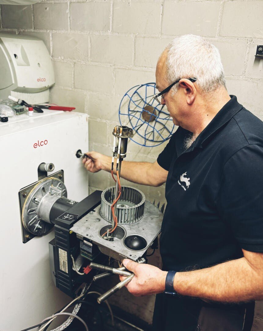 Ein Mann mit grauen Haaren und Brille repariert oder wartet eine Maschine, wobei er ein Teil mit Drähten und Werkzeugen neben einem an der Wand montierten Industriegerät mit der Aufschrift elco in einer Werkstatt hält.