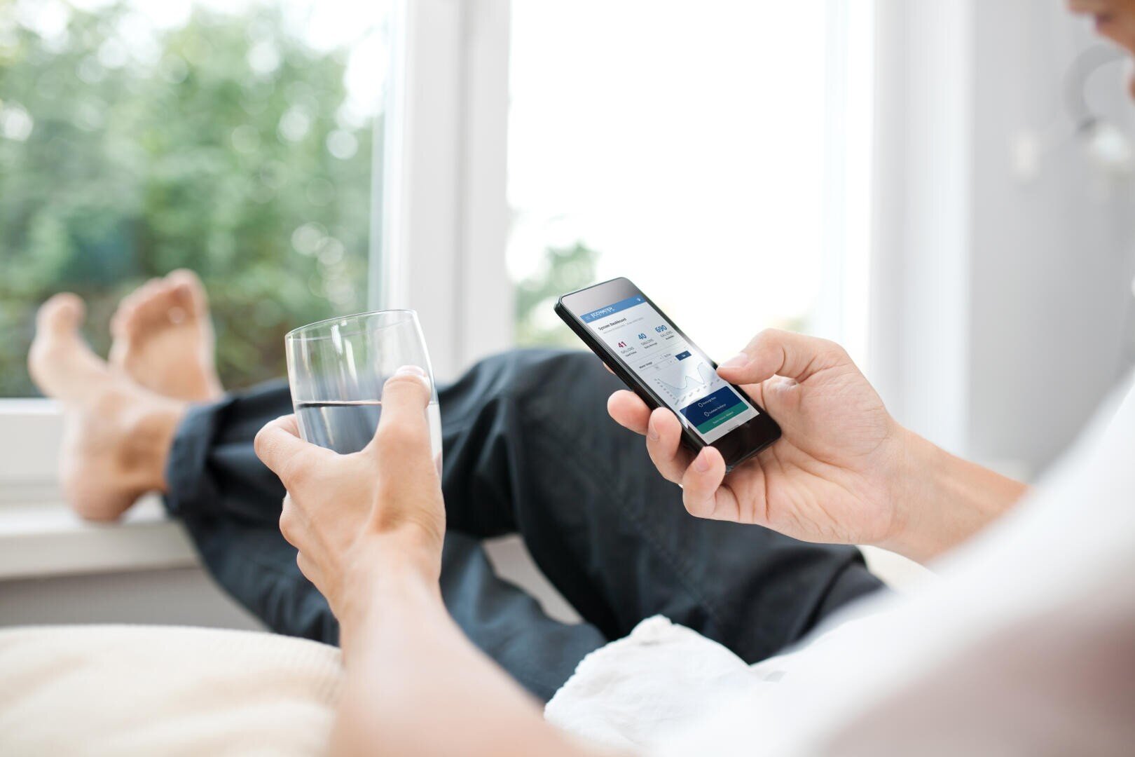 Eine Person entspannt sich in einem Innenraum am Fenster, hält ein Glas Wasser in der einen Hand und benutzt mit der anderen ein Smartphone, um auf einer Website oder einer App zu surfen. Die Beine sind bequem übereinander geschlagen.