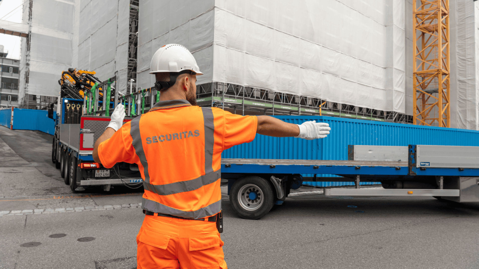 Un ouvrier du bâtiment en tenue de sécurité et casque orange dirige un camion qui recule sur un chantier, avec des échafaudages et des matériaux de construction visibles à l'arrière-plan.