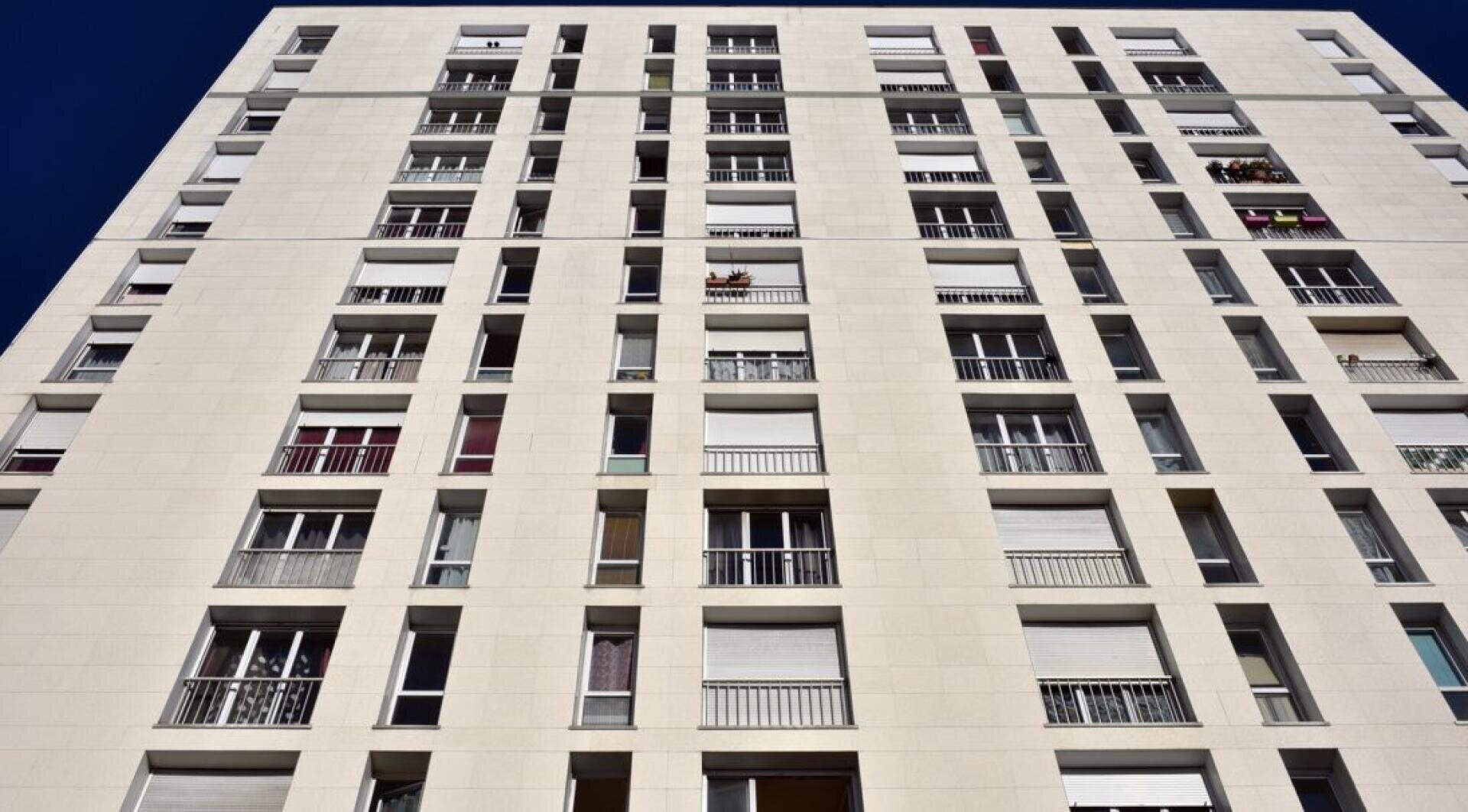Un immeuble d'habitation haut et moderne avec une façade blanche, de nombreuses fenêtres et plusieurs balcons sur lesquels se trouvent des plantes ou des objets. La photo est prise d'en bas, avec une vue sur l'immeuble dans un ciel bleu et dégagé.