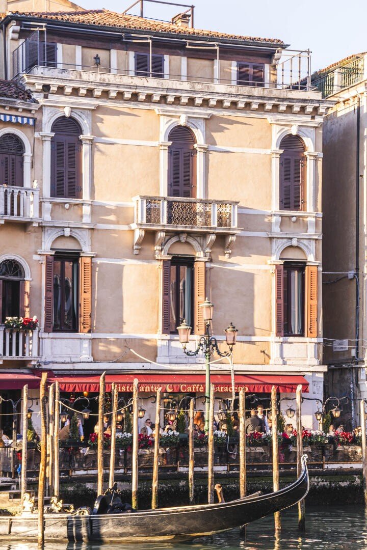 Ein sonniges dreistöckiges venezianisches Gebäude mit Bogenfenstern und hölzernen Fensterläden steht am Ufer eines Kanals. Gondelpfähle und eine traditionelle Gondel befinden sich im Vordergrund, zusammen mit einer Restaurantterrasse am Wasser.