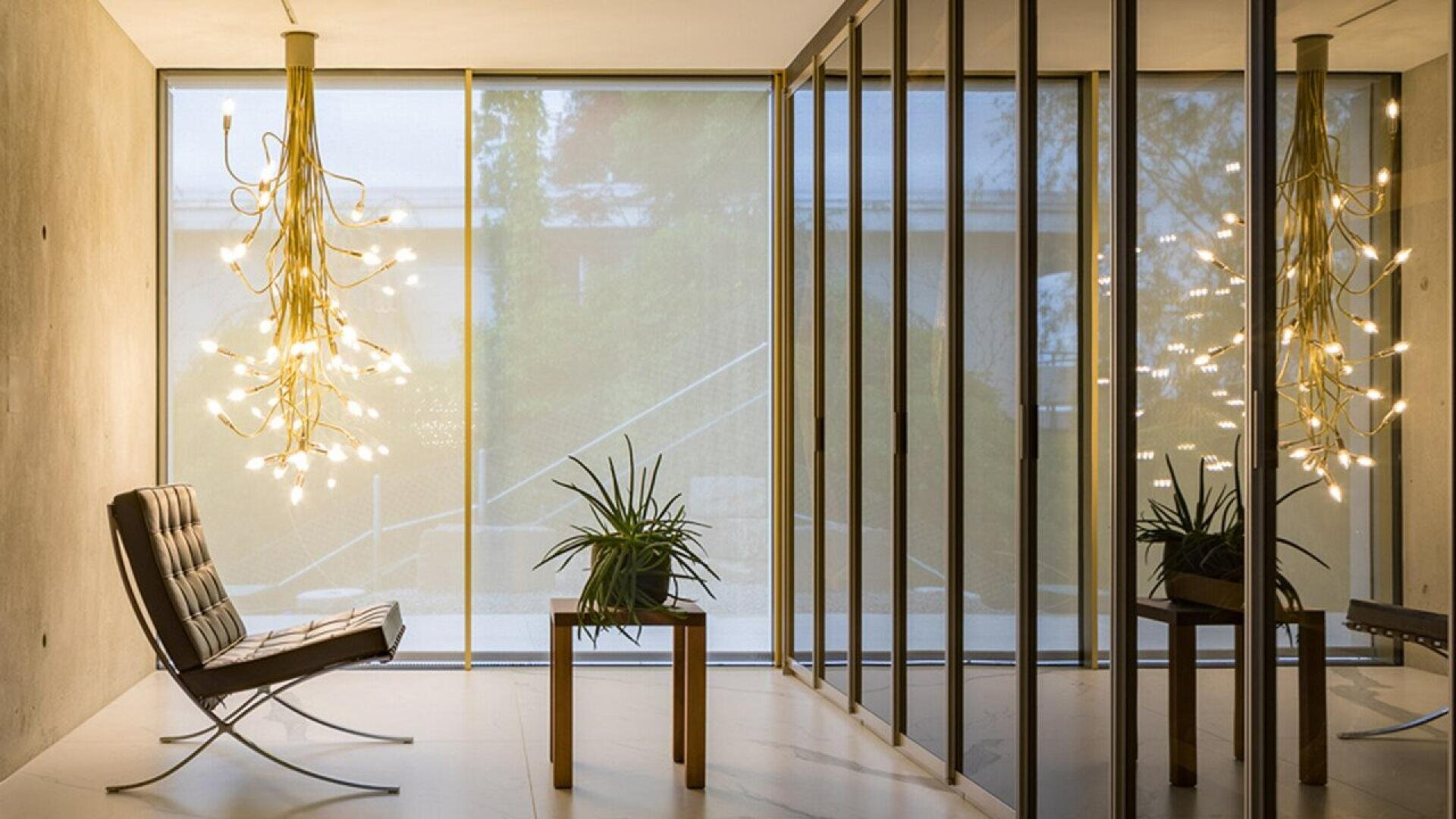 Intérieur moderne avec un fauteuil Barcelona en cuir capitonné, une plante sur une table d'appoint en bois et un lustre doré spectaculaire avec de nombreuses ampoules.