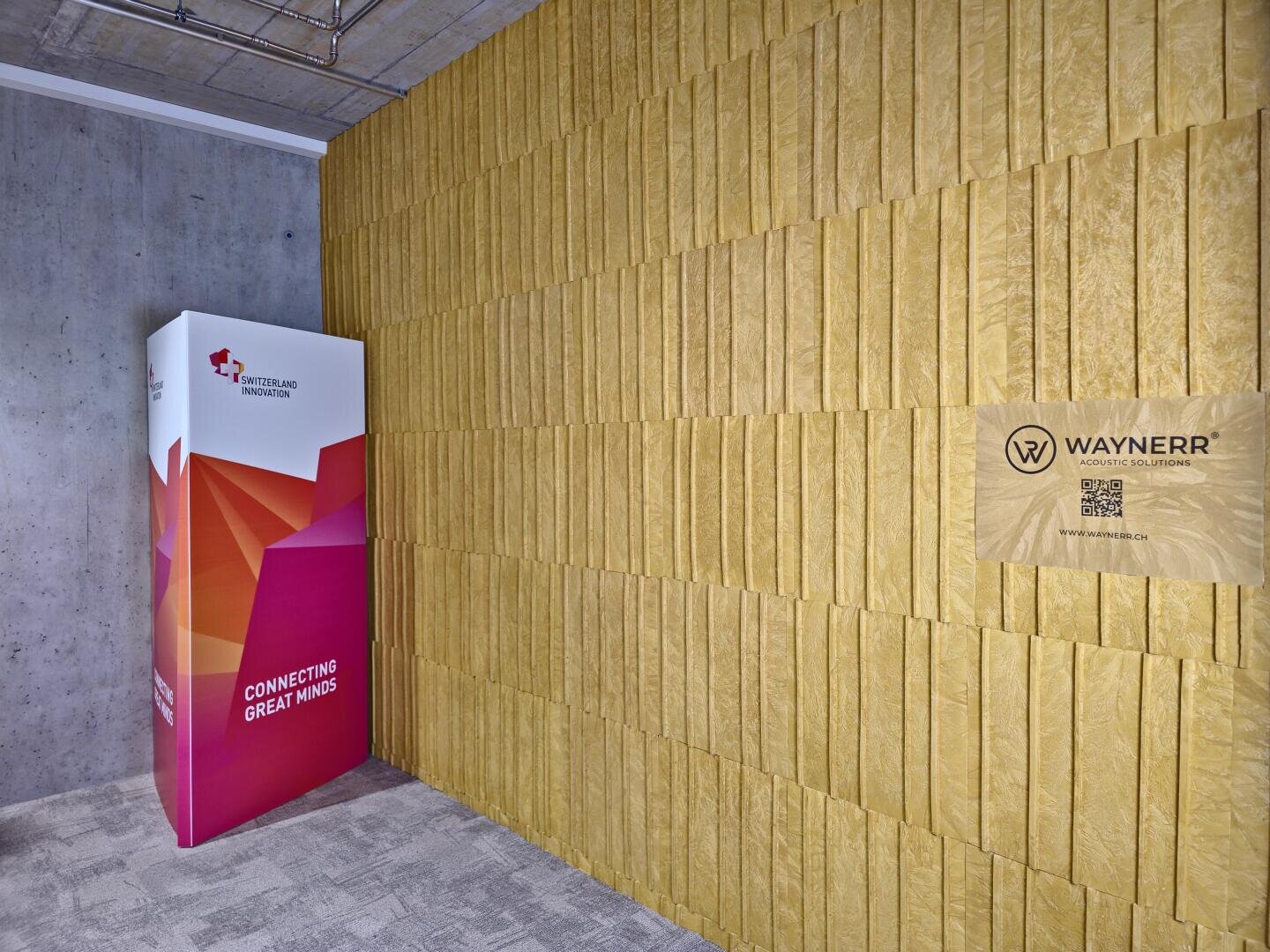 Un coin de bureau moderne présente un mur doré structuré avec un panneau Waynerr Robotics Solutions et un code QR, à côté d'un présentoir Switzerland Innovation au design géométrique rouge avec les mots "Connecting Great Minds".