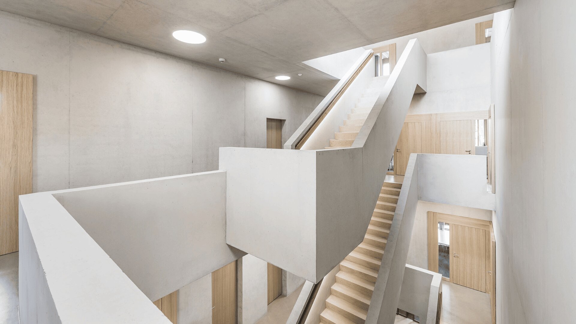 Un intérieur moderne et minimaliste avec des murs et des plafonds en béton, des couloirs ouverts et des portes en bois clair. Des escaliers géométriques centraux relient plusieurs étages, créant des lignes épurées et des angles vifs sous la lumière du jour et du plafond.