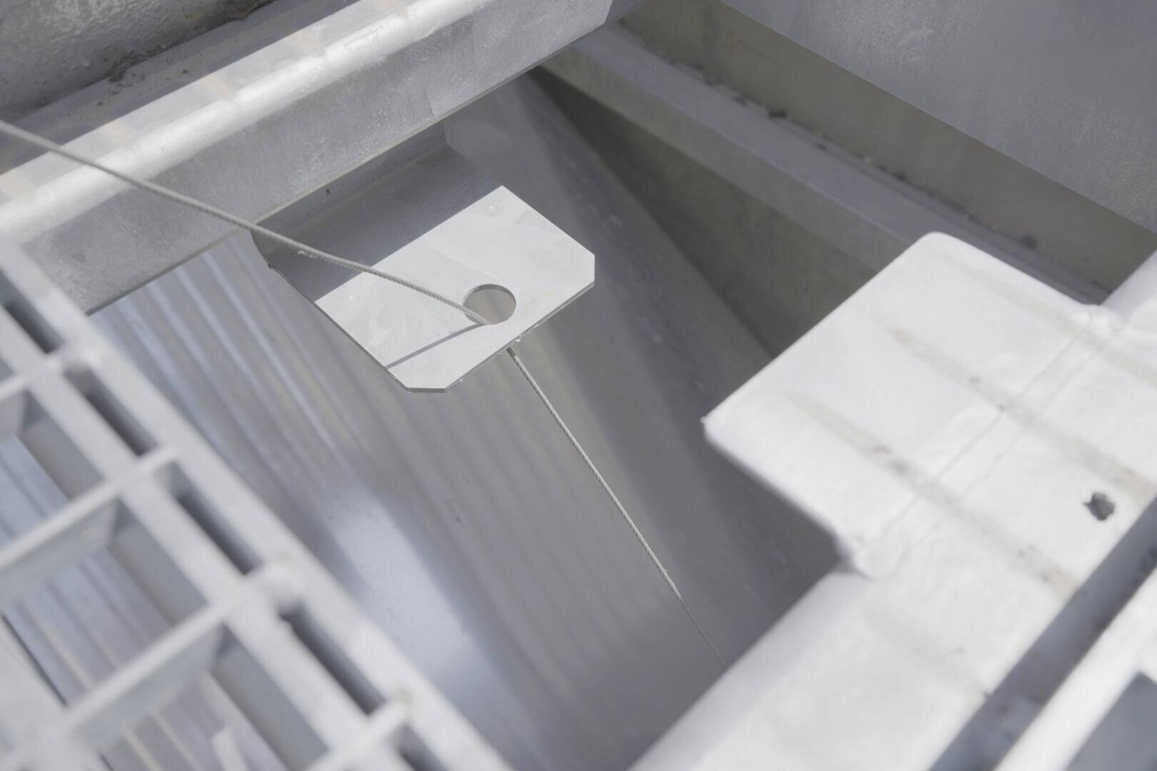 Nahaufnahme einer industriellen Metallstruktur mit einer vergitterten Plattform, einer geriffelten Oberfläche und einer Metallplatte mit einem runden Loch, die durch einen gespannten Draht oder ein Kabel verbunden ist.