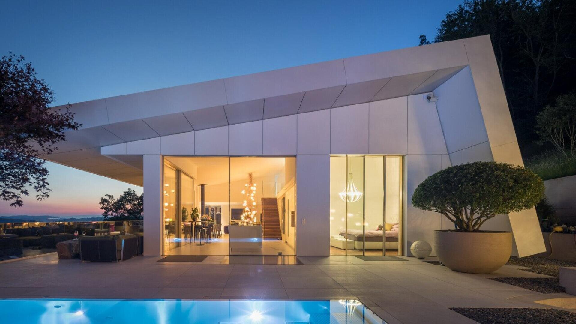Maison moderne blanche avec piscine au crépuscule. Les lumières intérieures éclairent la salle à manger, l'escalier et la chambre à coucher.