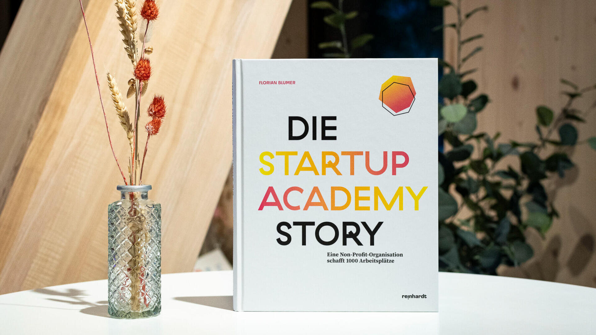 Ein Buch mit dem Titel Die Startup Academy Story steht aufrecht auf einer weißen Fläche neben einer Glasvase mit Trockenblumen, hinter denen ein hölzerner Hintergrund zu sehen ist.