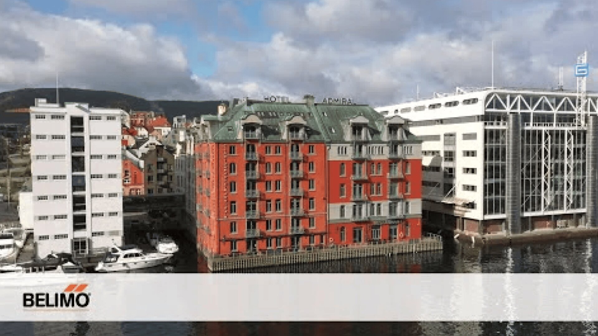 Eine Szene am Wasser zeigt ein rotes historisches Gebäude mit der Aufschrift Hotel Ambra zwischen modernen weißen Gebäuden, mit angedockten Booten in der Nähe, unter einem teilweise bewölkten Himmel. Das BELIMO-Logo erscheint in der unteren linken Ecke.