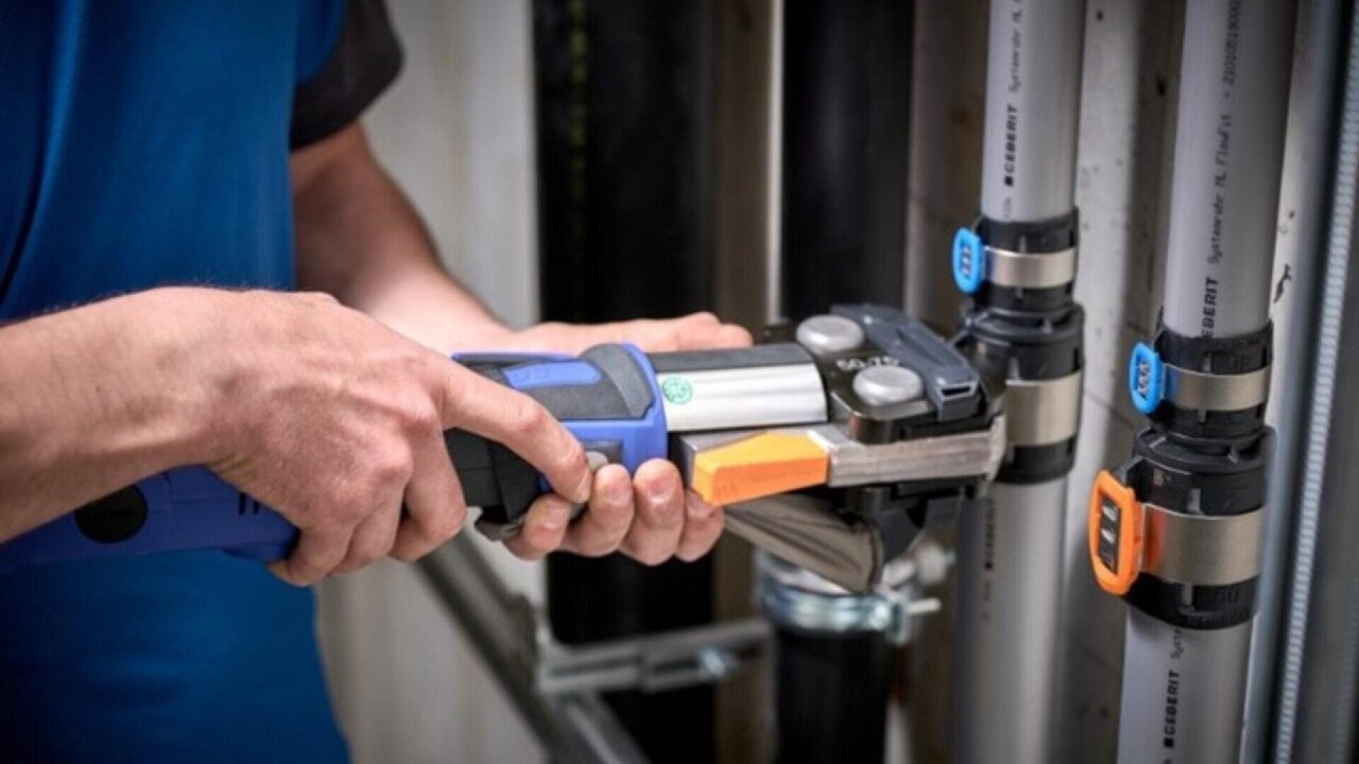 Une personne utilise un outil électrique manuel bleu et noir pour fixer des raccords sur des tuyaux en PVC gris fixés à un mur, lors de travaux de plomberie ou d'installation.