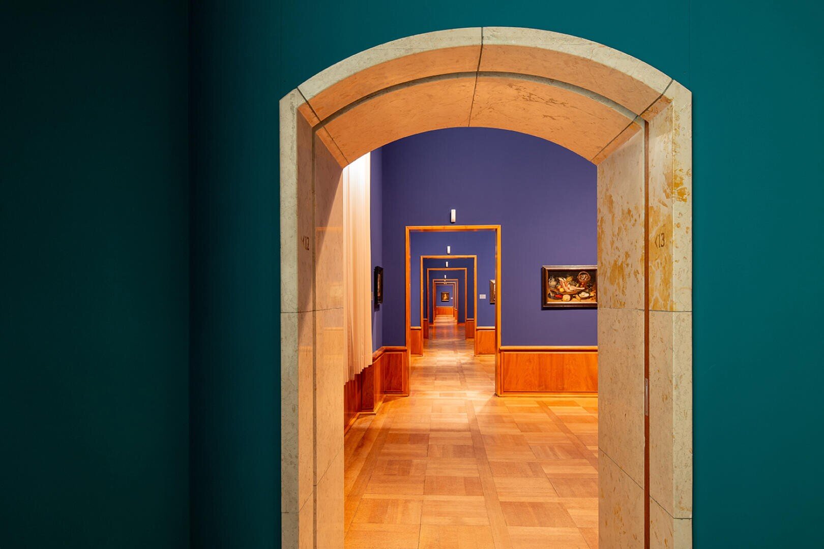 Une série de portes voûtées dans un musée ou une galerie, encadrant la suivante dans un long couloir au sol en bois, aux murs bleu-vert et bleu et aux tableaux suspendus le long des côtés.