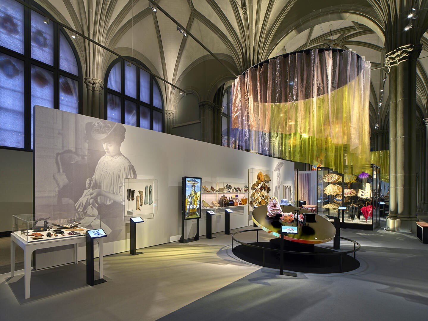 Eine moderne Museumsausstellung mit beleuchteten Displays von Accessoires und Hüten, interaktiven Bildschirmen, einem großen Vintage-Foto einer Frau und einer farbenfrohen, durchscheinenden Stoffinstallation, die in einer großen Halle von der Decke hängt.