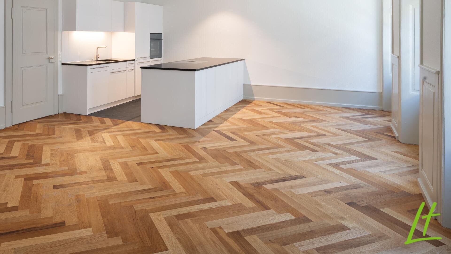 Cuisine moderne avec des armoires blanches et un îlot central qui se transforme en une pièce spacieuse avec un parquet en bois à chevrons. La pièce est bien éclairée par la lumière naturelle provenant de hautes fenêtres.