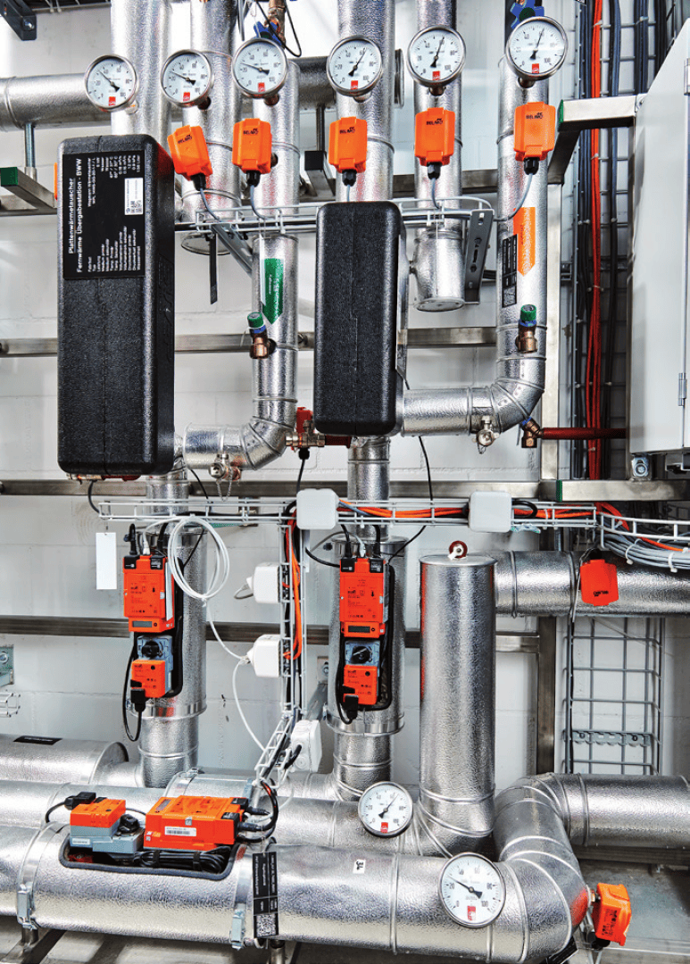 Un réseau complexe de tuyaux métalliques isolés avec des manomètres, des vannes orange et des boîtiers de commande, faisant probablement partie d'un système industriel de chauffage ou de climatisation, monté sur un mur blanc.