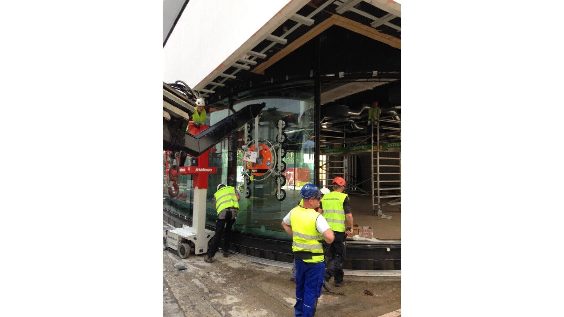 Des ouvriers du bâtiment portant des gilets de sécurité et des casques installent un grand panneau de verre incurvé à l'aide d'un élévateur à succion et d'une grue sur un chantier moderne avec échafaudage et poutres de plafond apparentes.