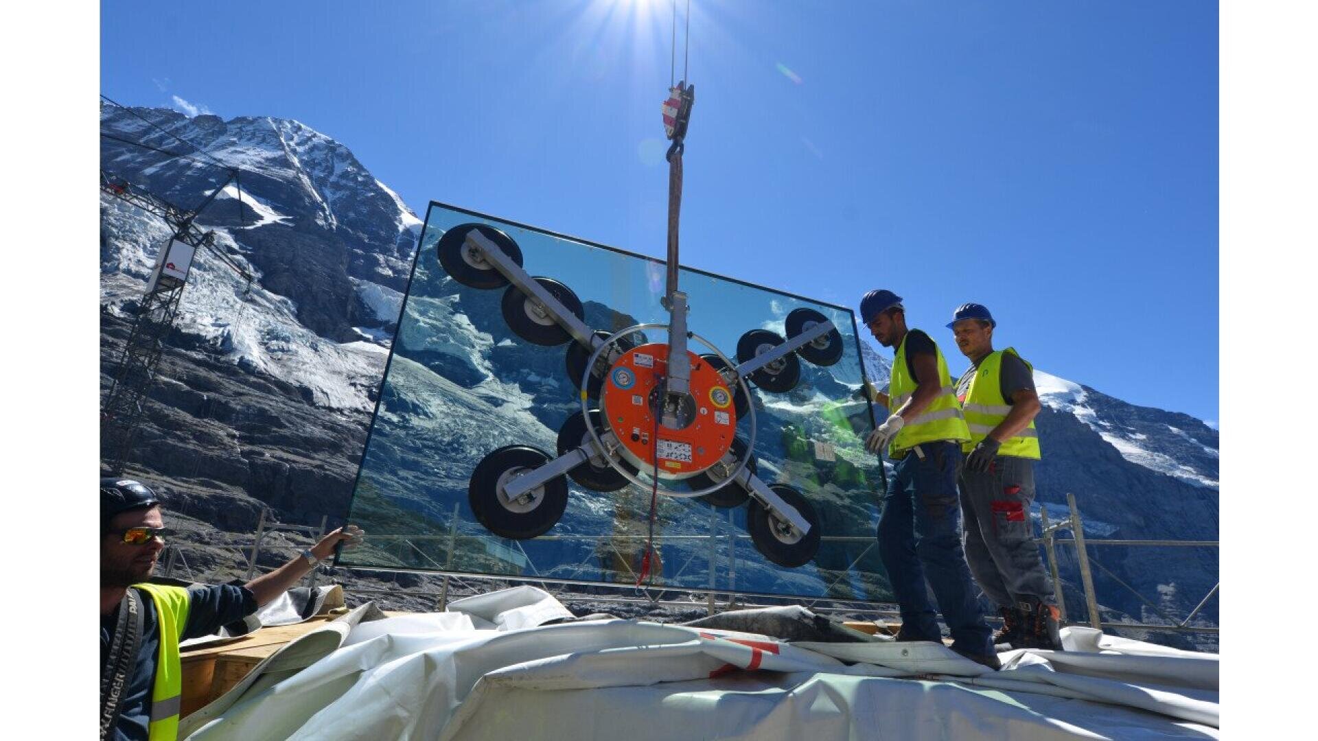 Trois ouvriers du bâtiment portant des équipements de sécurité guident un grand panneau de verre soulevé par une grue, avec des montagnes enneigées en arrière-plan et un ciel bleu clair au-dessus.