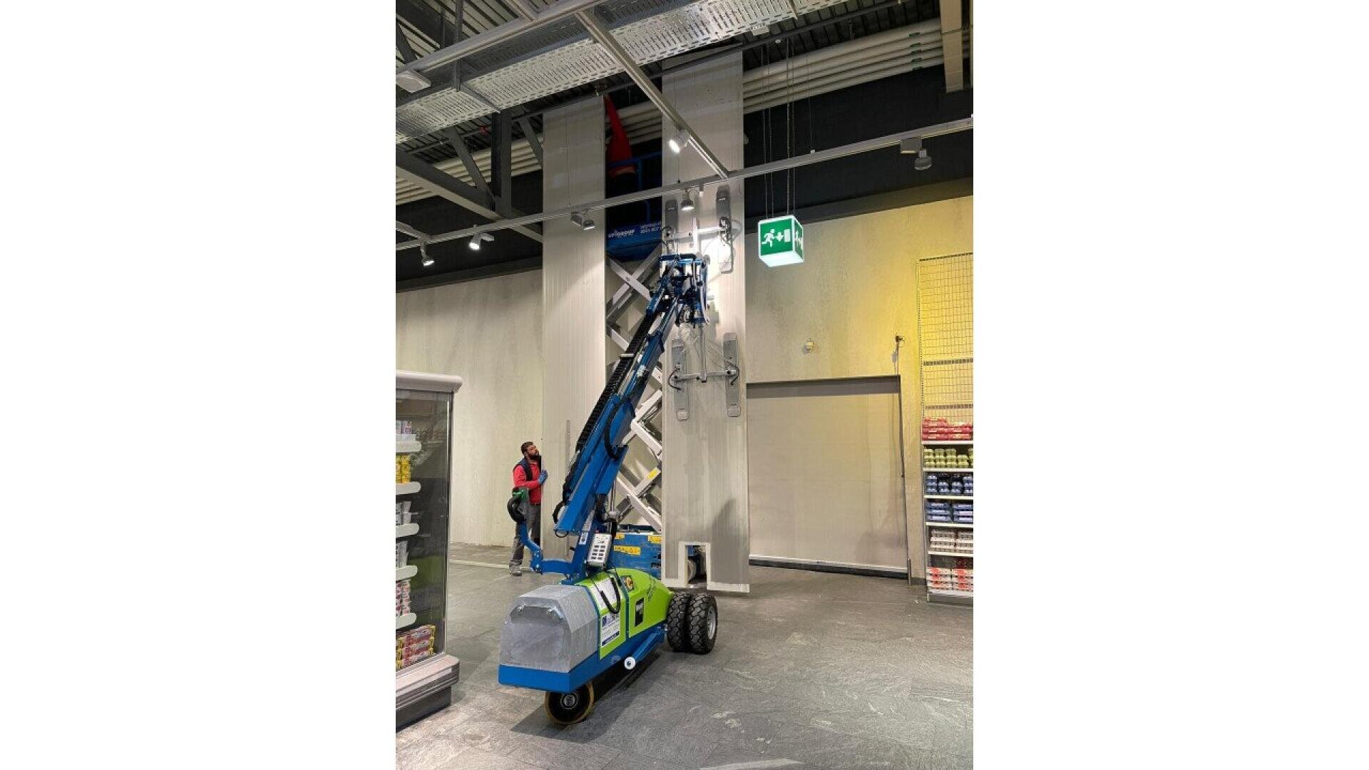 Un élévateur à ciseaux bleu et vert est levé vers le plafond d'un magasin, avec un employé à proximité et des étagères de produits de part et d'autre. Un panneau de sortie vert est visible au-dessus de l'élévateur.