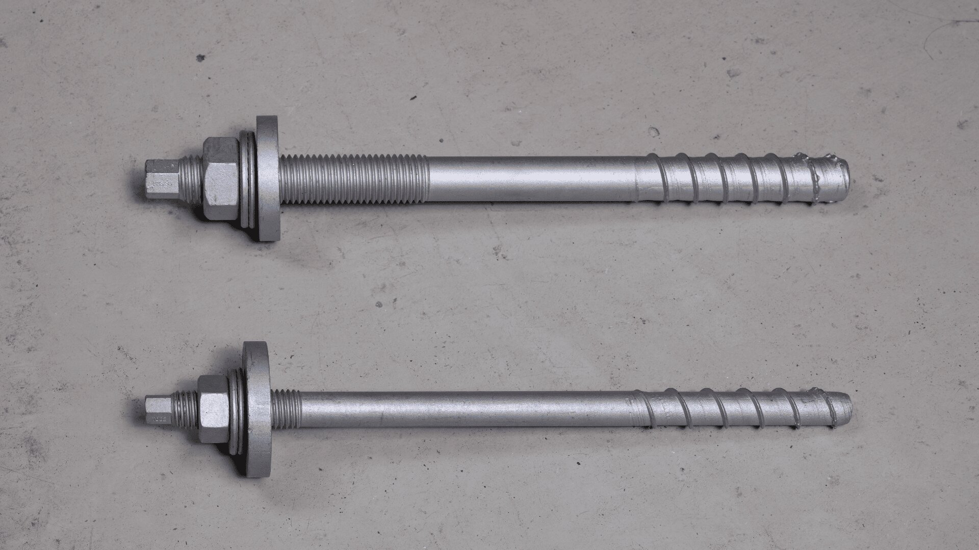 Zwei metallene Ankerbolzen mit Unterlegscheiben und Muttern liegen parallel auf einer hellgrauen Fläche, einer länger als der andere. Beide Bolzen haben einen Gewindeteil und einen gerillten Teil.