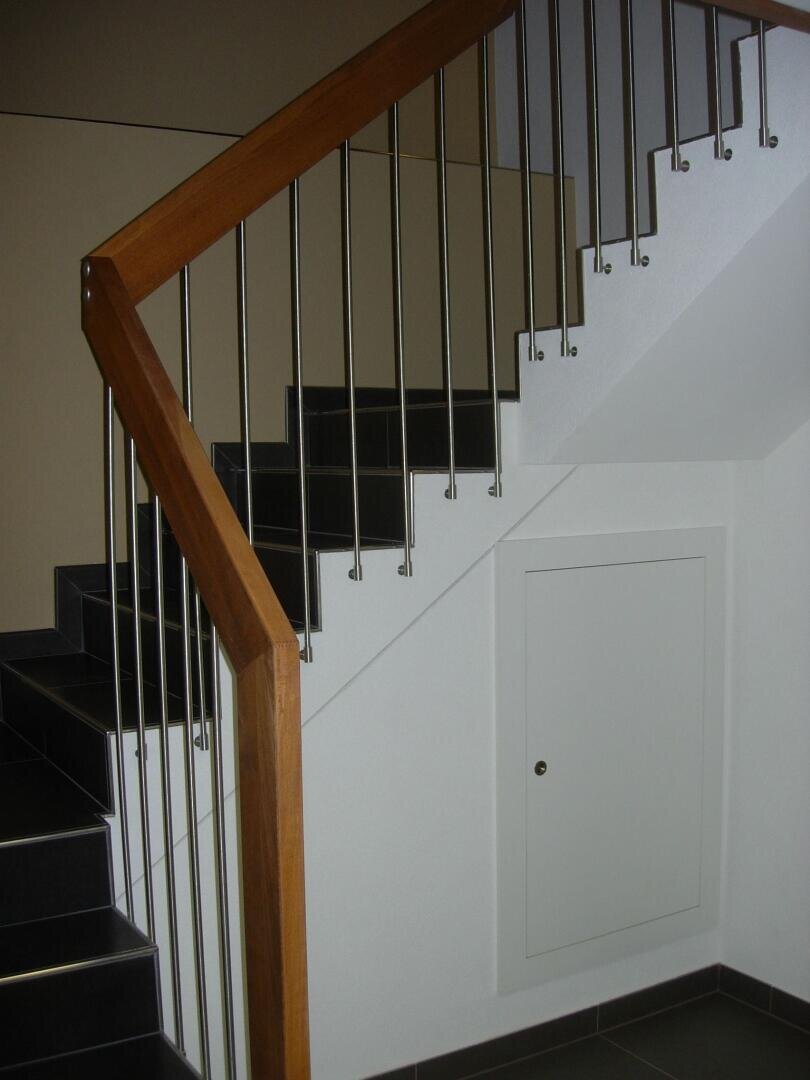 Eine moderne Innentreppe mit schwarzen Stufen, Metallgeländer und einem Holzhandlauf. Unter der Treppe befindet sich eine kleine weiße Tür, die in die Wand eingelassen ist.