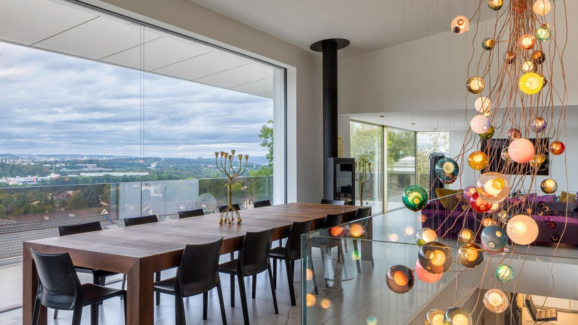Une salle à manger moderne avec une longue table en bois, des chaises noires et une grande fenêtre donnant sur l'horizon de la ville. Un lustre contemporain avec des orbes de verre colorés est suspendu au premier plan.
