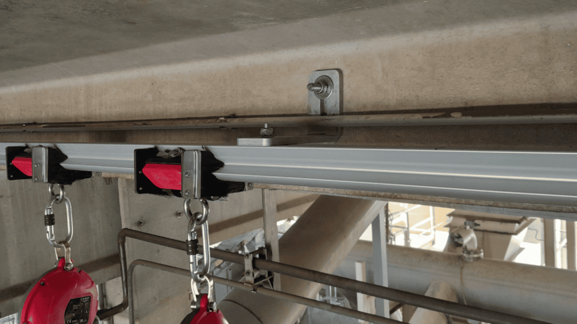 Gros plan d'un système de rail métallique aérien auquel sont attachées deux lignes de vie rouges à enrouleur pour la protection contre les chutes, montées sur une structure en béton dans un environnement industriel.
