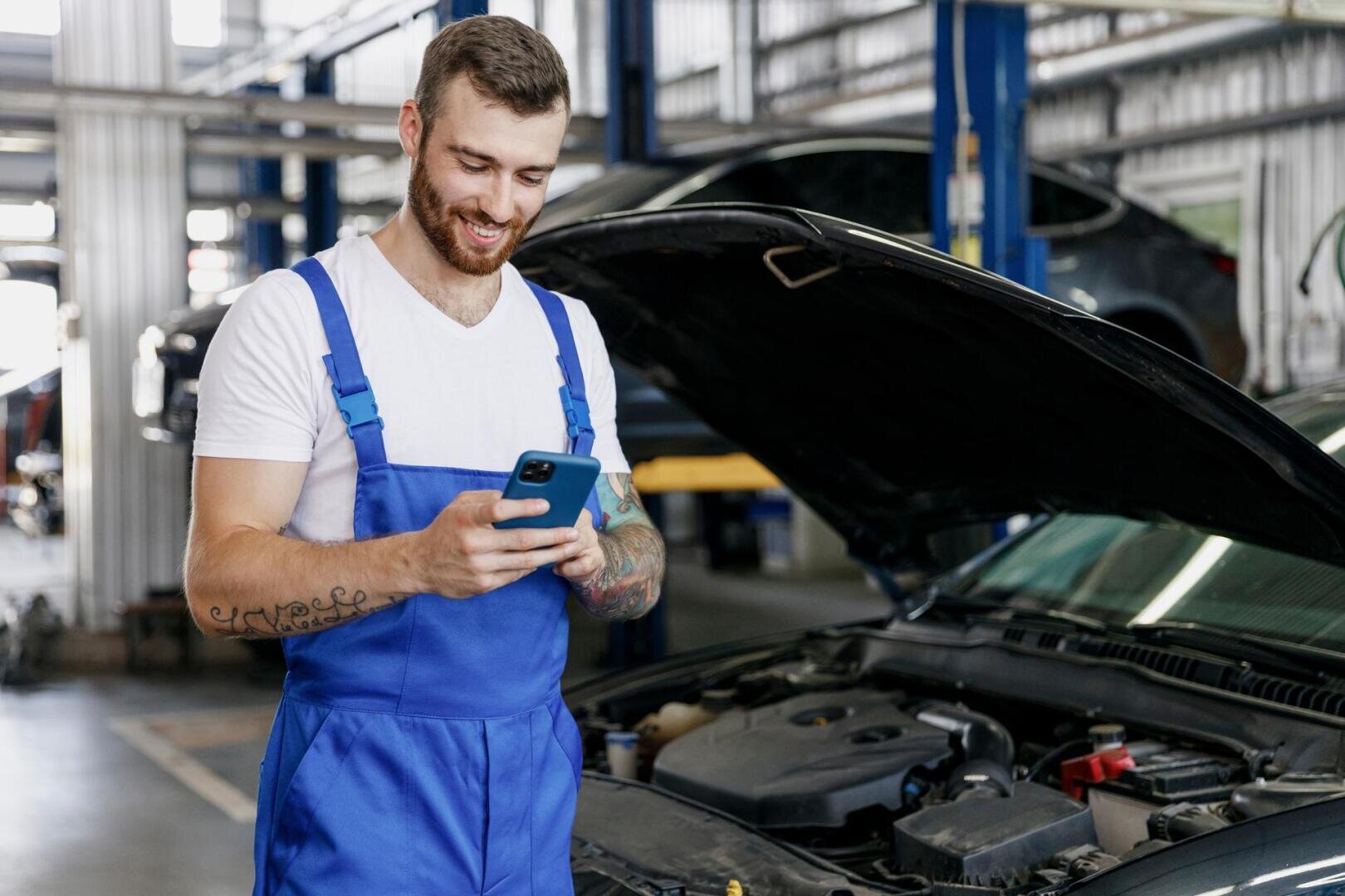 Un mécanicien souriant, vêtu d'une combinaison bleue, se tient à côté d'une voiture au capot ouvert et regarde un smartphone dans un atelier de réparation automobile.