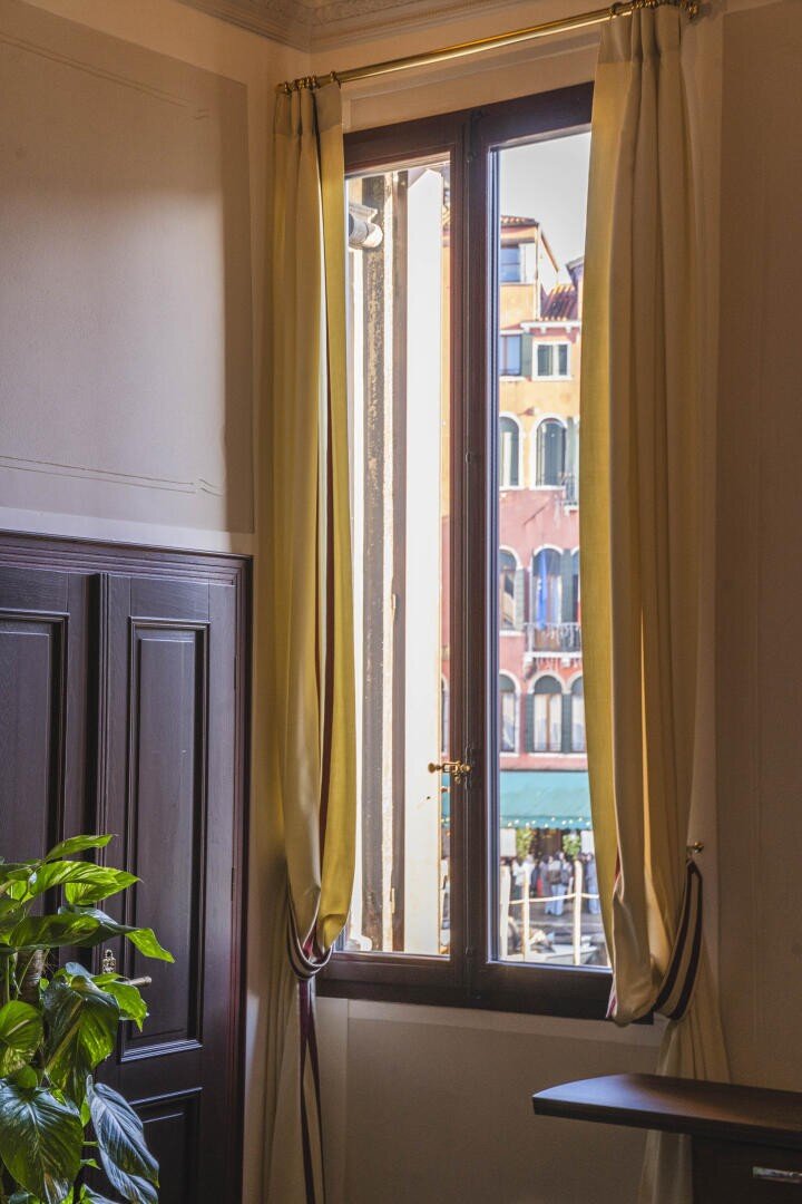 Das Sonnenlicht fällt durch ein Fenster mit offenen gelben Vorhängen und gibt den Blick auf bunte Gebäude im Freien frei. Im Inneren des Raumes sind ein Teil eines Holzmöbels und eine grüne Topfpflanze zu sehen.