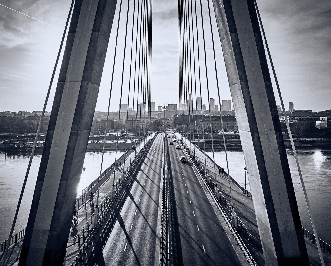 Eine Schwarz-Weiß-Ansicht einer Schrägseilbrücke mit Autos und Fußgängern, die einen Fluss in Richtung Stadtsilhouette unter einem klaren Himmel überspannt. Die Seile der Brücke bilden kühne vertikale Linien im Vordergrund.