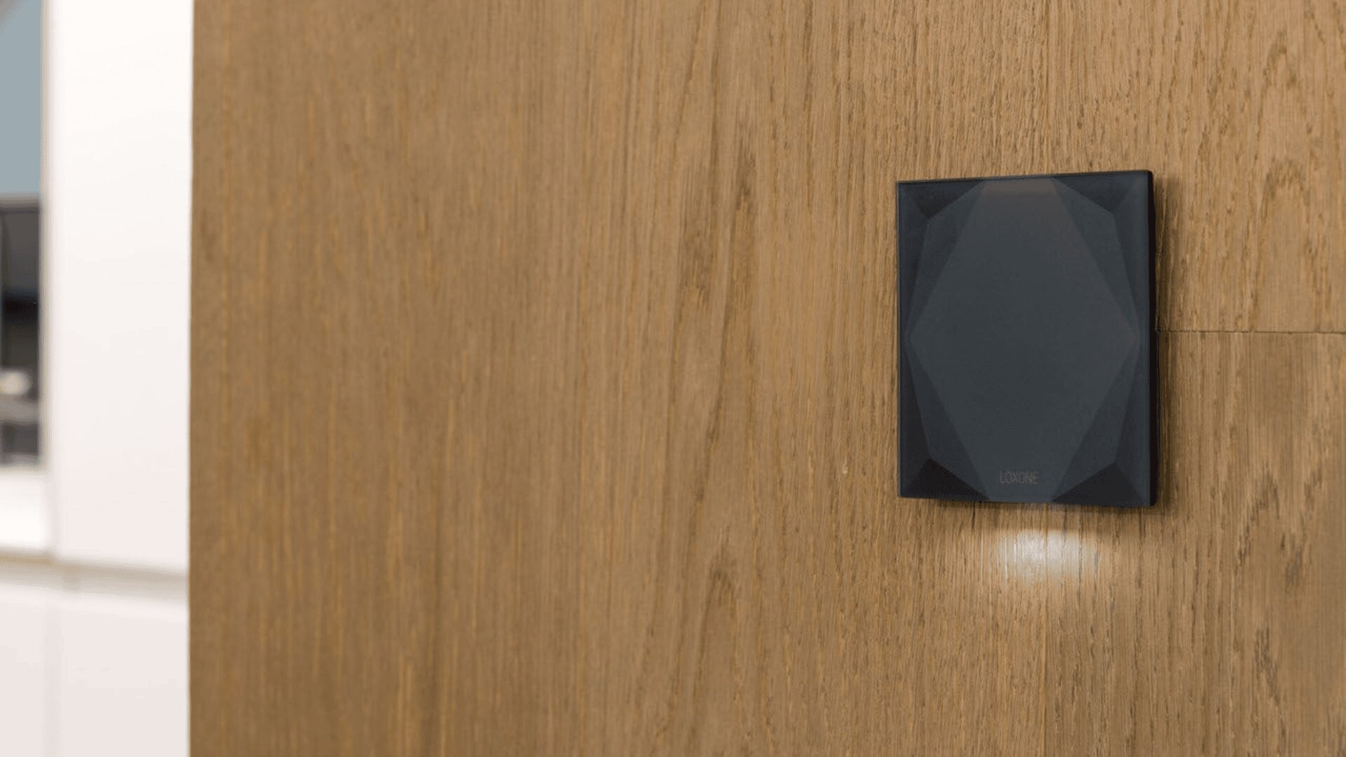 Ein schwarzes, geometrisches Wandgerät ist an einer hellen Holzwand befestigt. Das Gerät hat eine facettierte Oberfläche und eine kleine Kontrollleuchte darunter. Der Hintergrund zeigt ein modernes Interieur mit verschwommenen weißen Schränken.