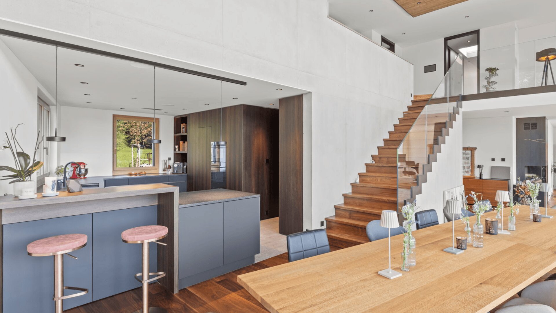 Moderner offener Küchen- und Essbereich mit blauen Schränken, Holzböden, Barhockern, einem Esstisch aus Holz mit Vasen und einer Treppe mit Glasgeländer, die in ein höheres Stockwerk führt. Der Raum ist von natürlichem Licht durchflutet.