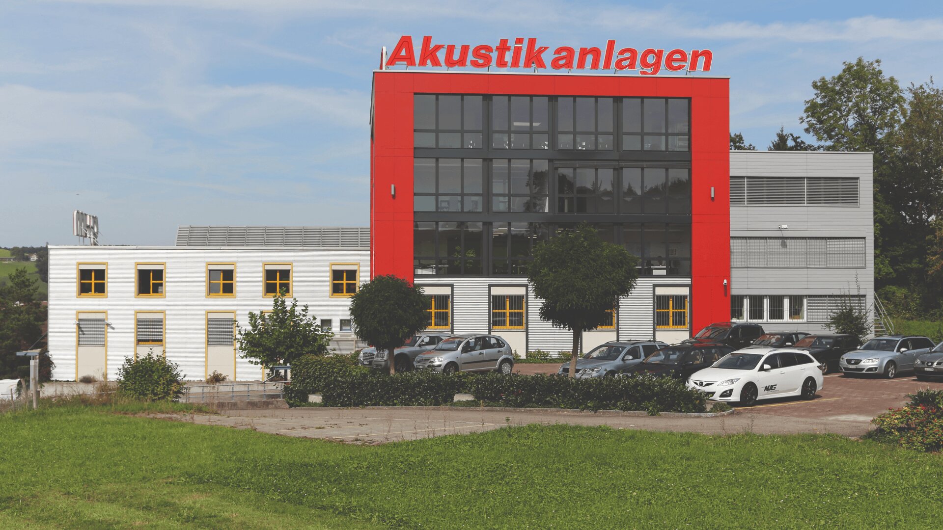 Ein modernes Gebäude mit einer roten Fassade, auf der in großen Buchstaben "Akustikanlagen" steht, umgeben von parkenden Autos, Grünflächen und Bäumen unter blauem Himmel.