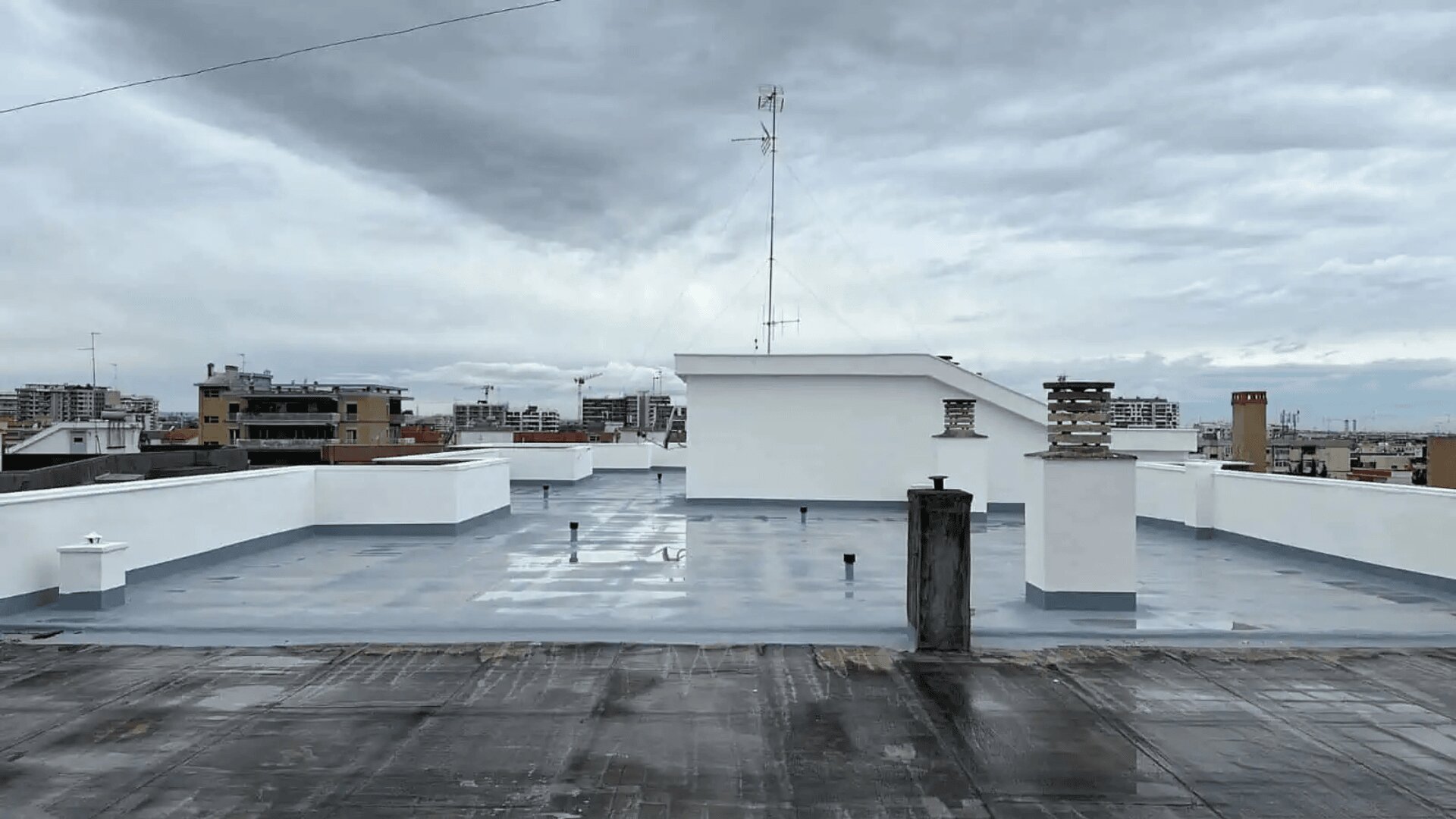 Un toit plat avec une surface lisse et grise, des tuyaux de ventilation et une structure blanche sous un ciel nuageux, avec des bâtiments urbains visibles en arrière-plan.