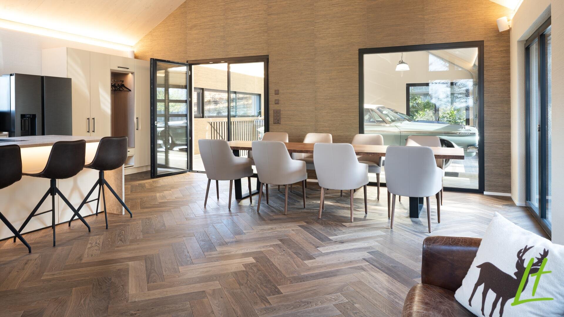 Salle à manger moderne avec des chaises claires autour d'une table en bois, un sol en bois à chevrons et de grandes fenêtres montrant une voiture à l'extérieur. Une cuisine avec des tabourets noirs et une chaise brune avec un coussin en forme de cerf sont également visibles.