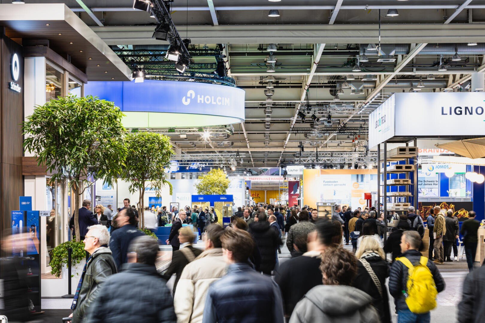 Un salon intérieur très animé avec de nombreuses personnes qui circulent entre les stands, y compris des stands d'entreprises comme Holcim. Un éclairage clair et des plantes vertes créent une atmosphère animée.