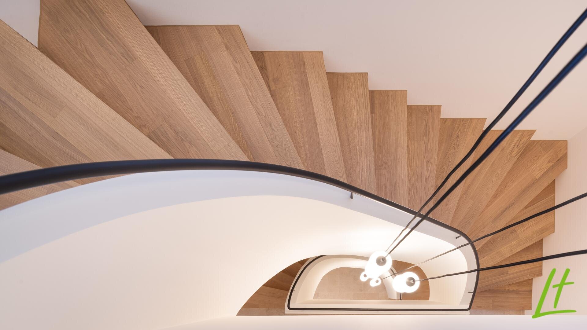 Vue de haut en bas d'un escalier en colimaçon moderne en bois, avec une rampe blanche incurvée et des lampes suspendues contemporaines accrochées au centre. Un logo vert lt est visible dans le coin inférieur droit.
