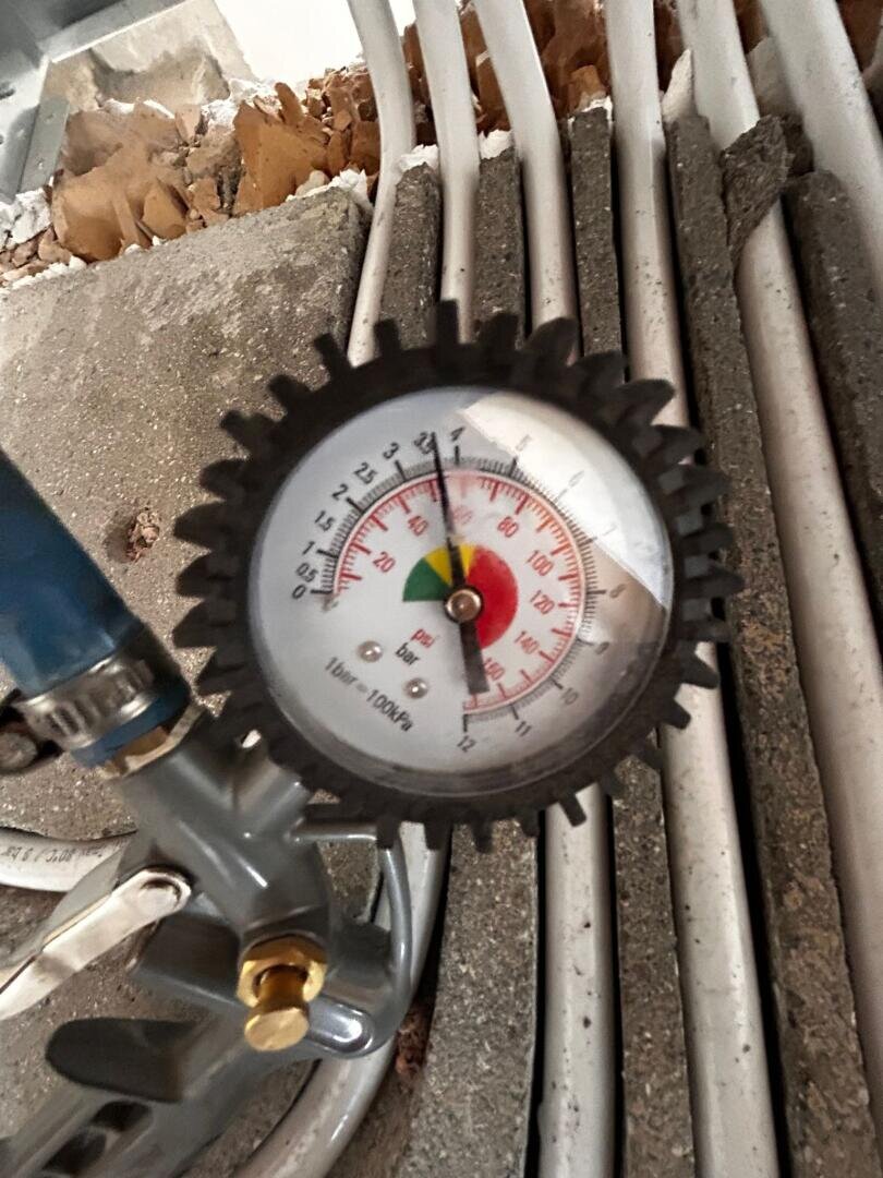Nahaufnahme eines Manometers mit einem schwarzen, zahnradähnlichen Rand, der etwa 2,4 bar anzeigt. Das Messgerät ist an Metallrohren befestigt, im Hintergrund sind mehrere weiße Kabel und Beton zu sehen.