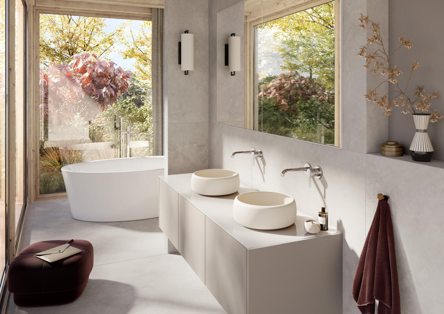 Modernes Badezimmer mit einer freistehenden weißen Badewanne an großen Fenstern, Doppelwaschbecken auf einem eleganten Waschtisch, Wandarmaturen und Herbstbäumen im Freien. Zur Dekoration gehören ein kastanienbraunes Handtuch, Keramikvasen und ein weicher Hocker.