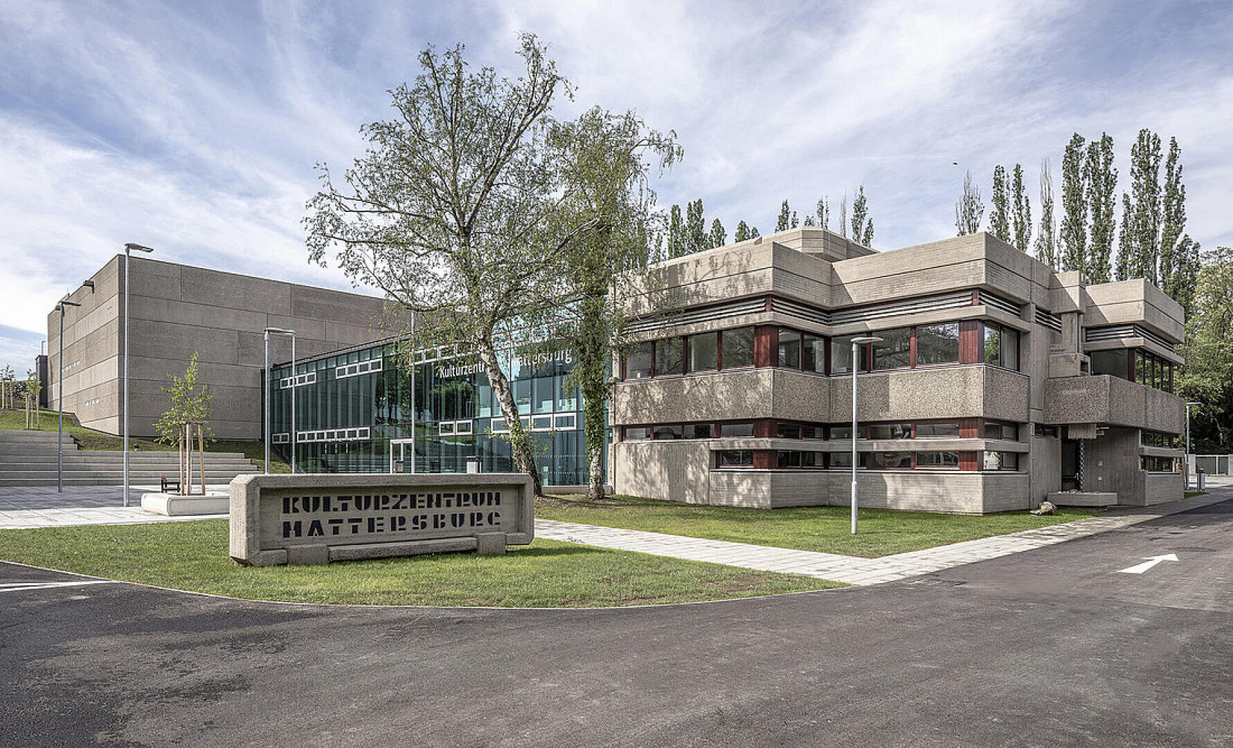 Moderner Gebäudekomplex aus Beton mit kantigen Formen und horizontalen Fenstern, umgeben von Bäumen und Gras. Davor steht ein Schild mit der Aufschrift Kulturzentrum Mattersburg. Der Himmel ist teilweise bewölkt.