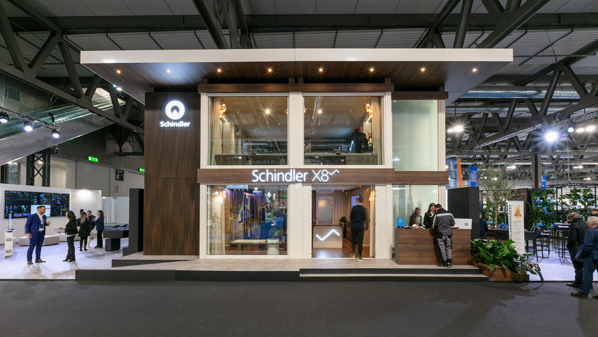 Stand d'exposition moderne sur deux étages pour Schindler, avec de grandes baies vitrées, des accents en bois, une signalétique éclairée et des personnes interagissant autour du stand dans un centre de congrès spacieux.