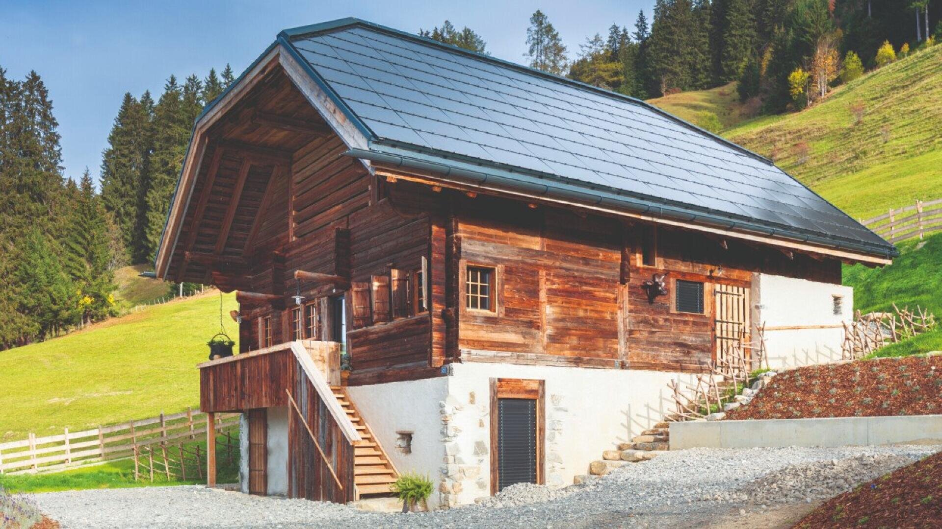 Une cabane rustique en bois avec un toit en pente équipé de panneaux solaires se trouve sur une colline herbeuse, entourée d'arbres et de clôtures en bois sous un ciel partiellement nuageux.