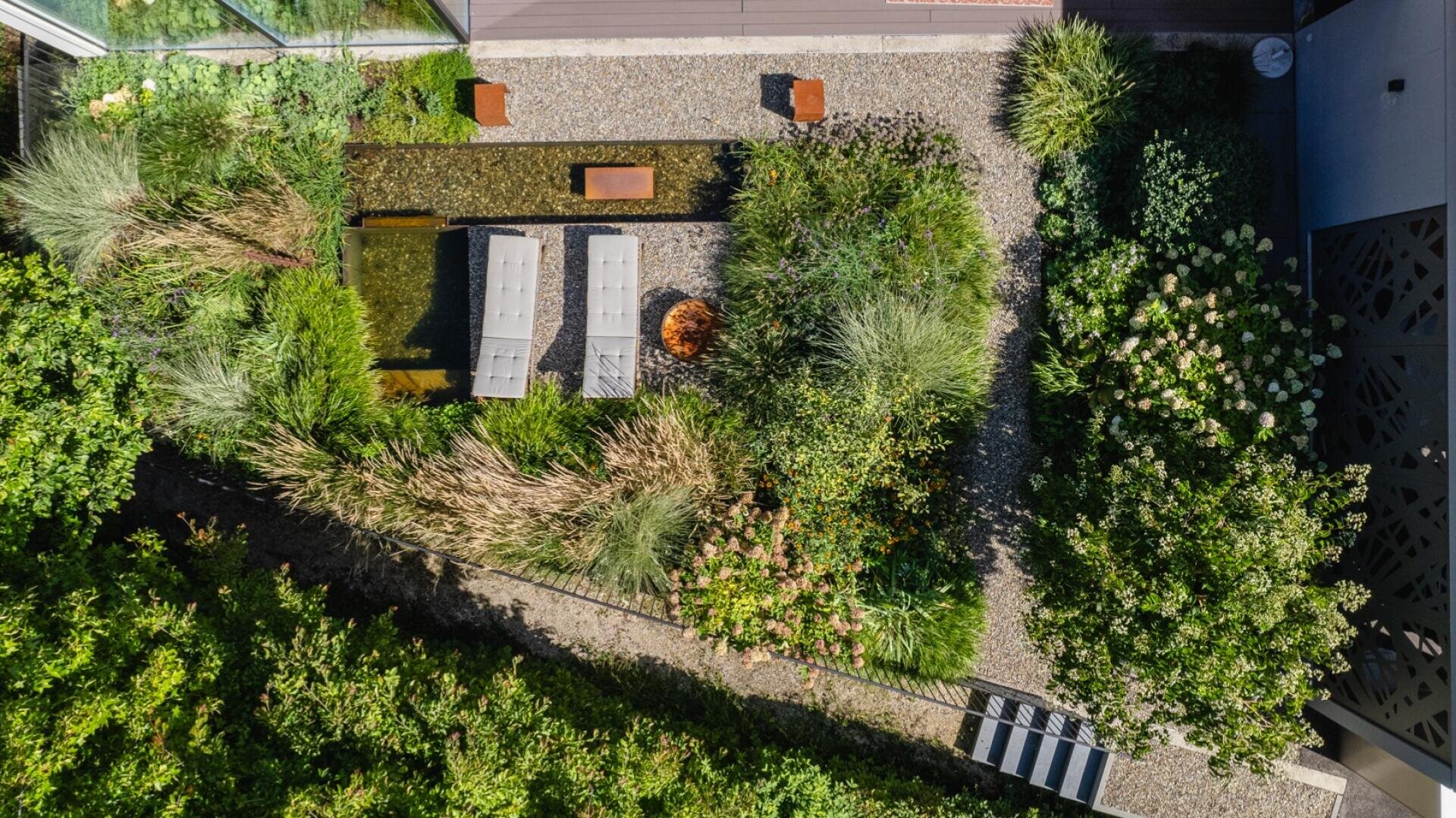Luftaufnahme eines modernen Gartens mit üppigen Grünpflanzen, Kieswegen, Bänken, einer Feuerstelle und einem schattigen Sitzbereich, angeordnet in einem geometrischen Design mit Wegen und natürlicher Landschaftsgestaltung.