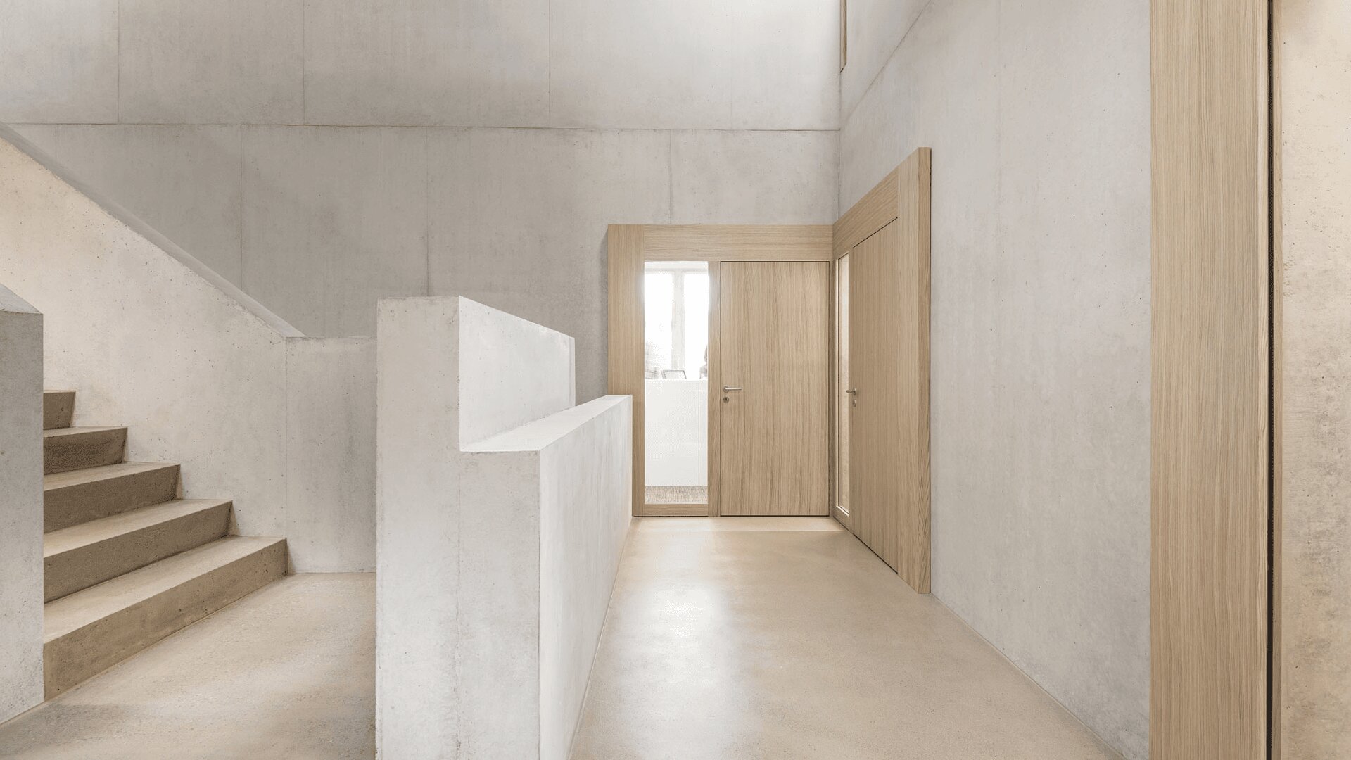Minimalistisches Interieur mit Wänden, Boden und Treppen aus beigem Beton. Türen und Holzverkleidungen aus hellem Holz kontrastieren mit den neutralen Farbtönen. Natürliches Licht dringt durch ein Fenster neben der Tür ein und schafft eine saubere, luftige und moderne Atmosphäre.