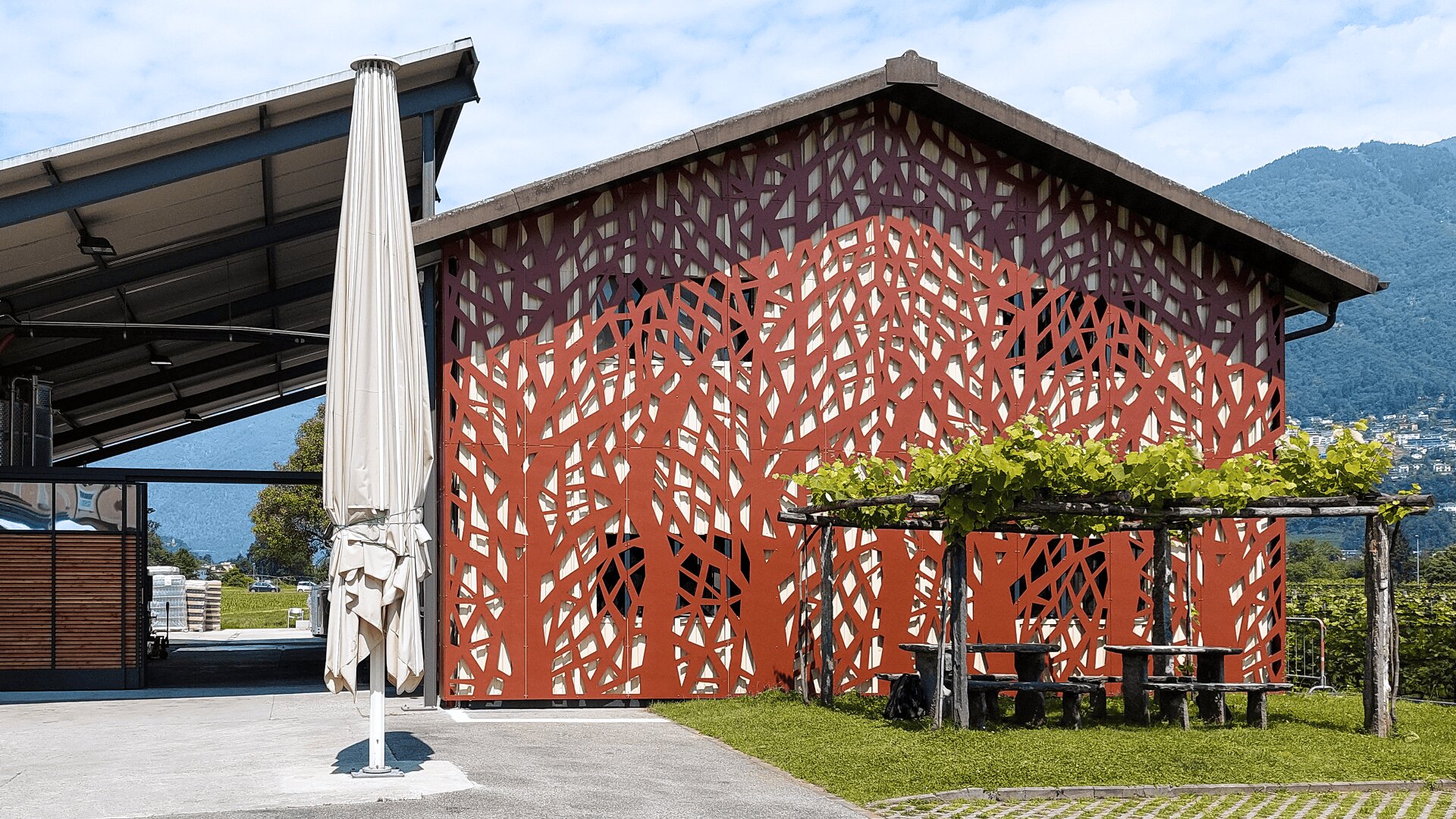 Ein Gebäude mit einer dekorativen, gitterartigen roten Fassade steht neben einem überdachten Außenbereich. Davor befinden sich ein schattiger Picknicktisch und ein aufrecht stehender, geschlossener Sonnenschirm. Im Hintergrund sind grüne Hügel und blauer Himmel zu sehen.
