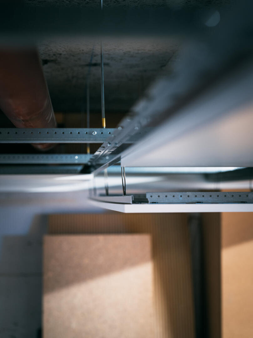 Nahaufnahme eines abgehängten Deckengerüsts mit Metallaufhängungen und -platten, von unten gesehen, mit unscharfen Baumaterialien und Rohren im Hintergrund.